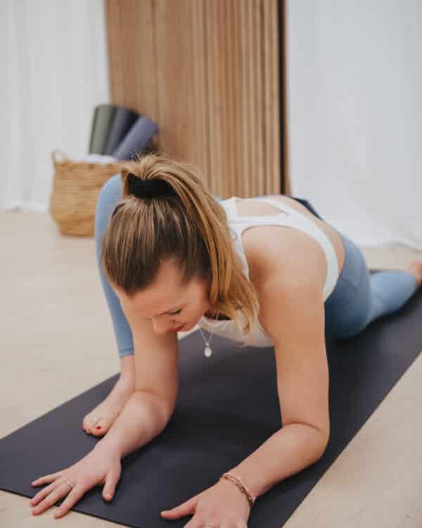 Een vrouw in een witte top en een blauwe legging die een yogastretch uitvoert op een donkere antislipmat in een minimalistische studio.