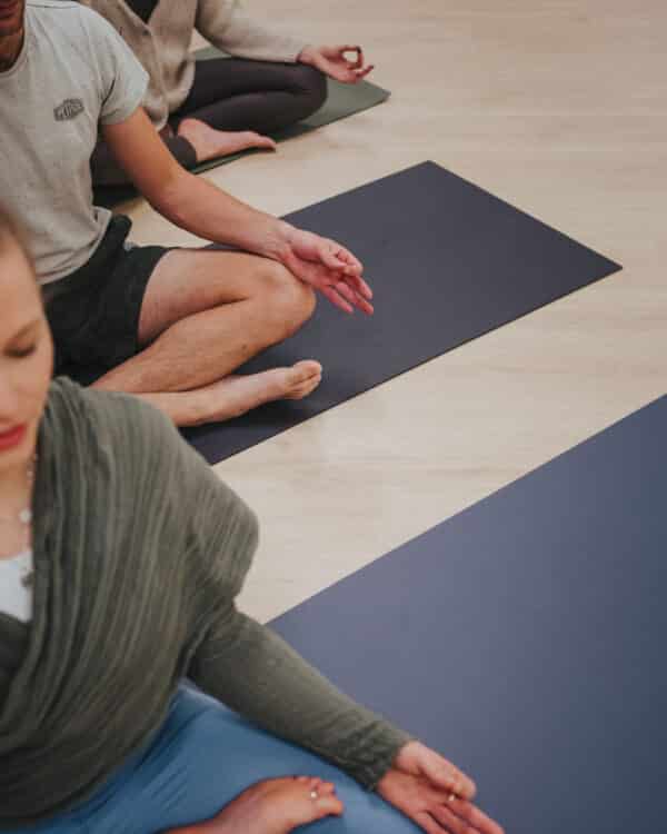 Top view of three people sitting in lotus pose on dark yoga mats during a group meditation class.