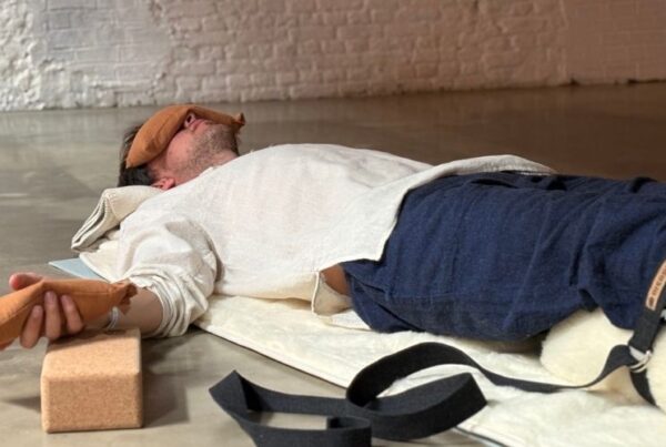 Yoga teacher assisting a student at ADM Antwerp studio alongside a practitioner resting in savasana on a Pierre Sports wool mat cover with cork block and yoga belt